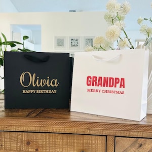 Puede incluir: Dos bolsas de regalo, una negra y otra blanca, sobre una superficie de madera. La bolsa negra tiene "Olivia" y "HAPPY BIRTHAY" en letras doradas. La bolsa blanca tiene "GRANDPA" y "MERRY CHRISTMAS" en rojo.