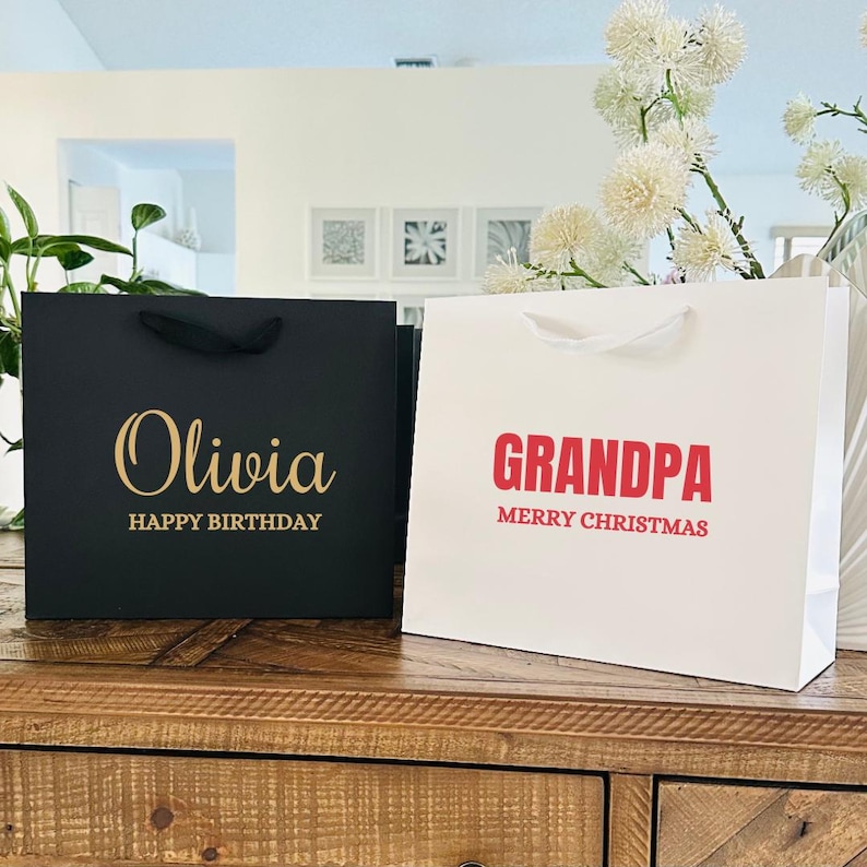 Puede incluir: Dos bolsas de regalo, una negra y otra blanca, sobre una superficie de madera. La bolsa negra tiene "Olivia" y "HAPPY BIRTHAY" en letras doradas. La bolsa blanca tiene "GRANDPA" y "MERRY CHRISTMAS" en rojo.