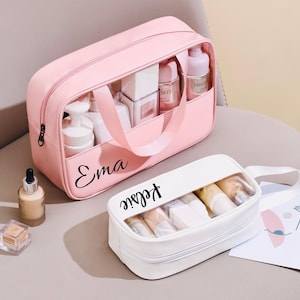 May include: Two pink and white travel makeup bags with clear see-through compartments. The pink bag has the name "Ema" written on it. The white bag has the name "Keelie" written on it.