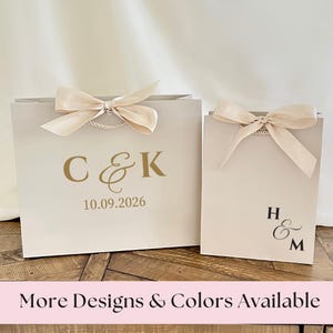 May include: Two beige gift bags with cream-colored bows. The larger bag has gold lettering with the initials "C & K" and the date "10.09.2026". The smaller bag has the initials "H & M". The text "More Designs & Colors Available" is at the bottom.