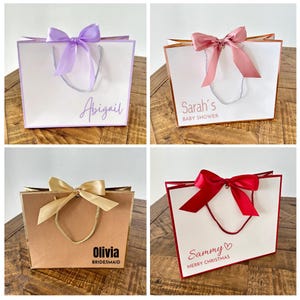 Pode incluir: Quatro sacos de presente com fitas de diferentes cores e mensagens personalizadas. Os sacos s&atilde;o brancos, castanhos e creme com guarni&ccedil;&otilde;es douradas e vermelhas. As mensagens nos sacos s&atilde;o "Abigail", "Ch&aacute; de Beb&ecirc; da Sarah", "Olivia Madrinha" e "Sammy Feliz Natal".