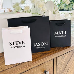 Puede incluir: Bolsas de regalo en blanco y negro con asas de sat&eacute;n blanco. Las bolsas tienen nombres impresos en una fuente negrita sin serifa. El texto incluye "STEVE TO MY GROOM", "JASON BEST MAN" y "MATT GROOMSMAN".