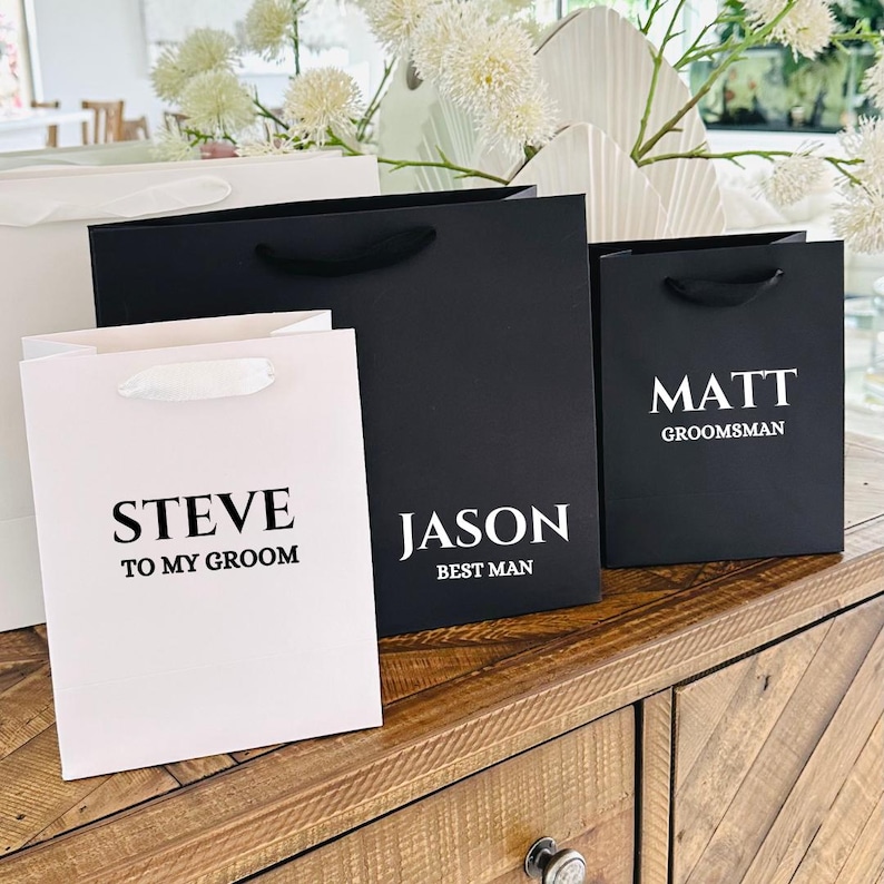 Puede incluir: Bolsas de regalo en blanco y negro con asas de sat&eacute;n blanco. Las bolsas tienen nombres impresos en una fuente negrita sin serifa. El texto incluye "STEVE TO MY GROOM", "JASON BEST MAN" y "MATT GROOMSMAN".