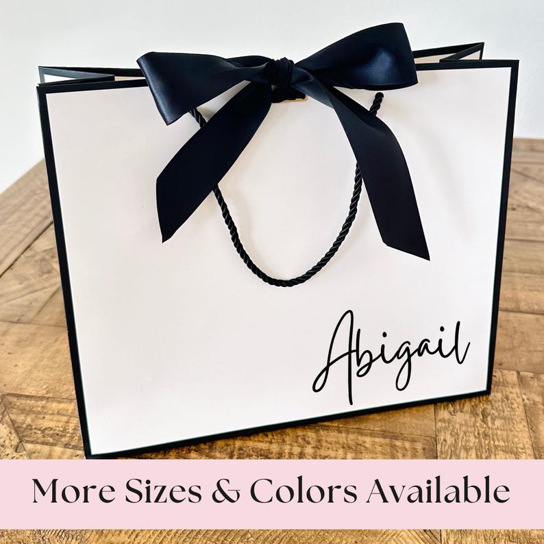 May include: White gift bag with a black ribbon and black handles. The bag has the name "Abigail" printed on the front.
