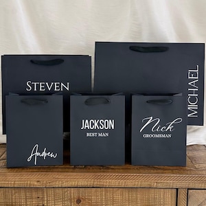May include: Five black gift bags with white text. The bags are arranged in a row on a wooden surface. The text on the bags reads: "Steven", "Andrew", "Jackson Best Man", "Nick Groomsman", and "Michael".