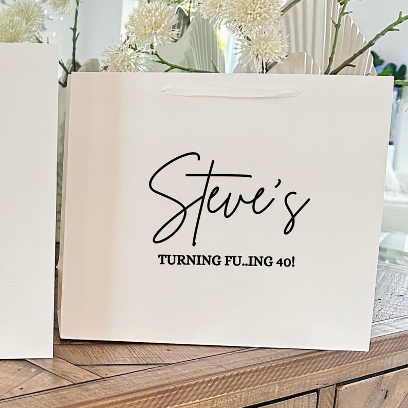 Puede incluir: Una bolsa de regalo blanca con el nombre "Steve's" en fuente cursiva y la frase "TURNING FU..ING 40!" impresa en negro. La bolsa tiene un asa de cinta blanca.