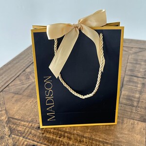 May include: A black gift bag with a gold border and gold rope handles. The bag has a gold bow and the word "MADISON" printed in gold on the front.