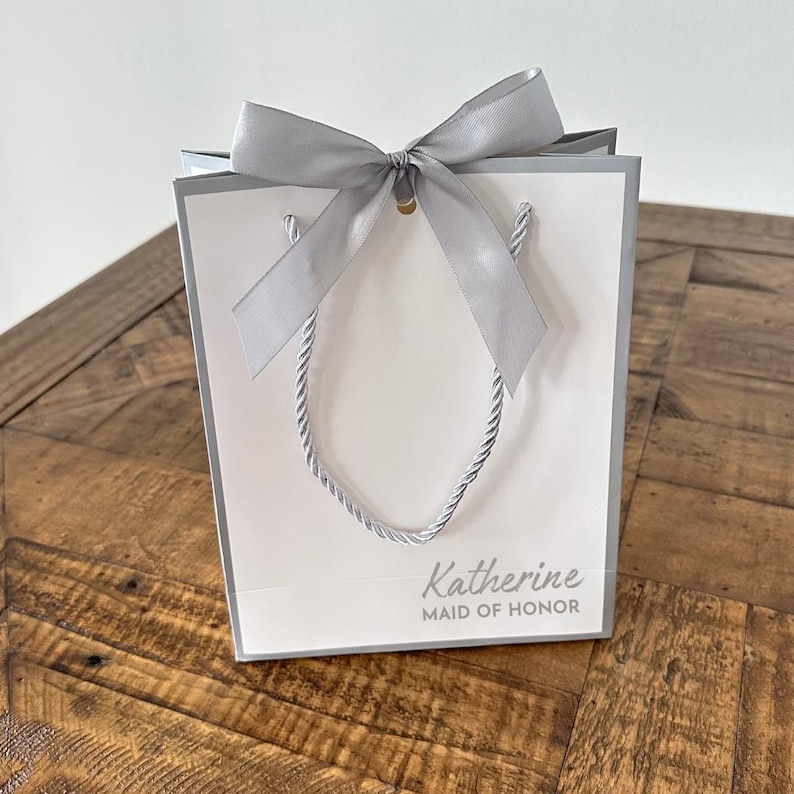 Pode incluir: Uma sacola de presente branca com um la&ccedil;o prateado e o texto "Katherine Maid of Honor" impresso na frente.