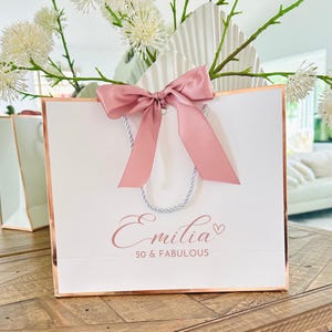 Pode incluir: Uma sacola de presente branca com uma borda de ouro rosa e um la&ccedil;o de cetim rosa. A sacola tem o texto "Emilia 50 & Fabulous" impresso na frente.