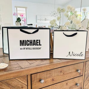 Puede incluir: Dos bolsas de regalo blancas con ribetes y asas negras sobre una superficie de madera. Una bolsa dice "MICHAEL 50 & STILL KICKIN'" en texto negro. La otra bolsa tiene el nombre "Nicole" en cursiva.
