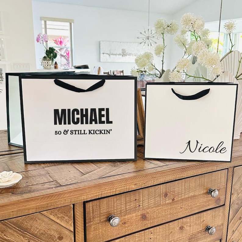 Puede incluir: Dos bolsas de regalo blancas con ribetes y asas negras sobre una superficie de madera. Una bolsa dice "MICHAEL 50 & STILL KICKIN'" en texto negro. La otra bolsa tiene el nombre "Nicole" en cursiva.