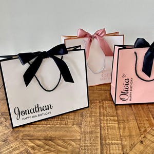 Pode incluir: Tr&ecirc;s sacolas de presente com la&ccedil;os de fita preta e texto personalizado. As sacolas s&atilde;o brancas, rosa e douradas. O texto nas sacolas diz "Jonathan Feliz anivers&aacute;rio de 40 anos", "Olivia Feliz anivers&aacute;rio" e "Venha celebrar o ch&aacute; de beb&ecirc;".