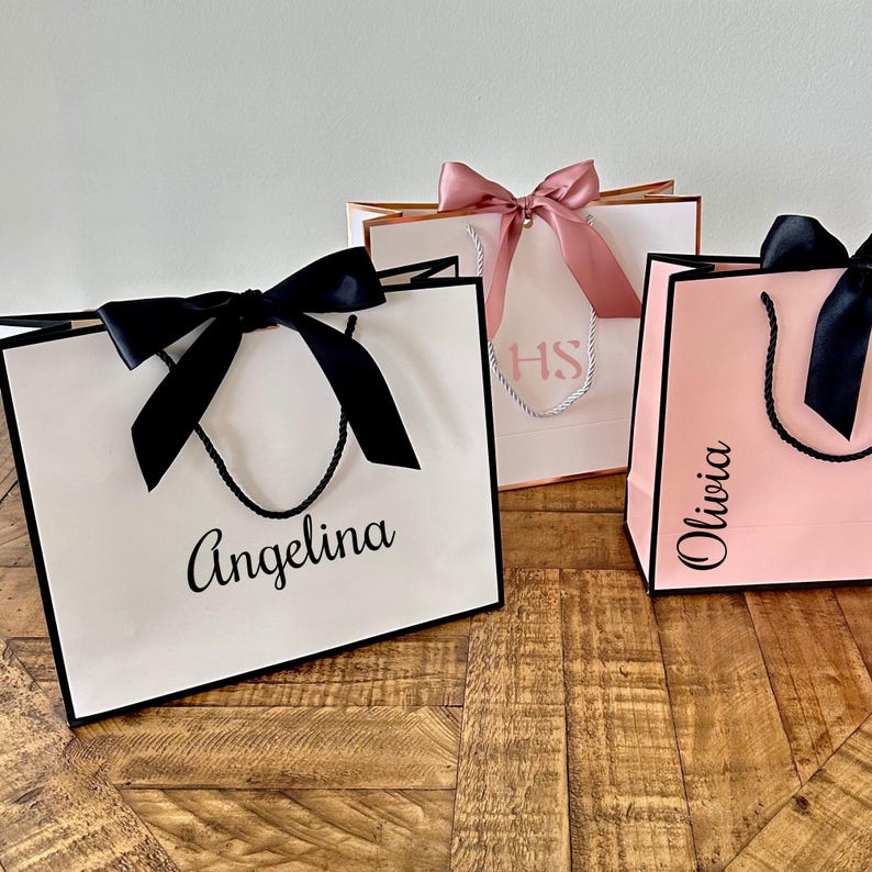 May include: Three white gift bags with black handles and black ribbon bows. The bags are personalized with the names "Angelina", "Olivia", and "HS".