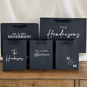 Pu&ograve; includere: Un gruppo di sacchetti regalo neri con testo bianco. Le borse presentano frasi come "MR. & MRS. HENDERSON", "The Hendersons" e le iniziali "H & M". Ogni borsa ha un manico nero.