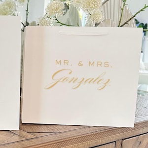Pu&ograve; includere: Una borsa regalo bianca con le parole "MR. & MRS." e il cognome "Gonzalez" in corsivo dorato. La borsa ha un manico a nastro bianco, adatta per un matrimonio o un evento speciale.