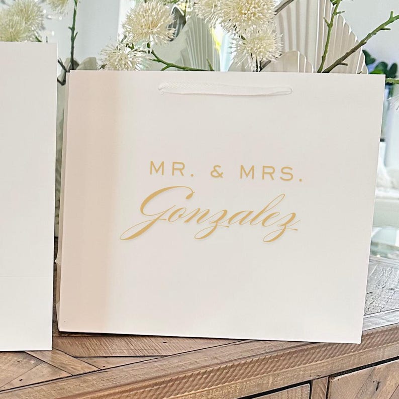 Pu&ograve; includere: Una borsa regalo bianca con le parole "MR. & MRS." e il cognome "Gonzalez" in corsivo dorato. La borsa ha un manico a nastro bianco, adatta per un matrimonio o un evento speciale.