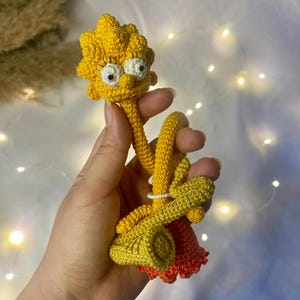 May include: A crocheted yellow and orange doll with a long neck and a long, thin body. The doll has a yellow head with a yellow and white face and black eyes. The doll is holding its arms in front of its body.