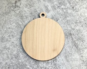 May include: A round, unfinished wooden ornament with a small hole at the top for hanging. The ornament is made of light-colored wood and has a smooth surface. The background is a mottled gray surface.
