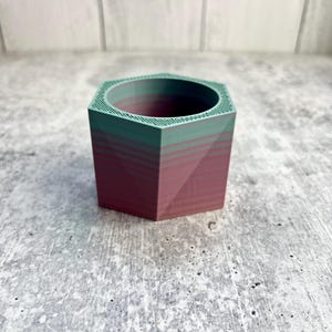 May include: A geometric planter with a hexagonal top and a square base. The planter is a gradient of colors, with a teal green top and a deep red-purple base. The planter is made of a solid material and is suitable for small plants.