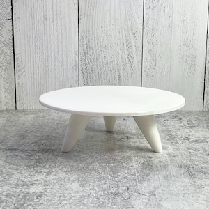 May include: A small, white, round table with three angled legs. The table is made of wood and has a smooth, clean surface. The design is minimalist and modern, suitable for displaying small items.