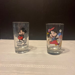 May include: Two clear glass drinking glasses with Mickey Mouse designs. The glass on the left has Mickey Mouse in red shorts and yellow shoes. The glass on the right has Mickey Mouse in a blue shirt and red shorts. Both glasses have the text "Mickey Mouse" printed on them.