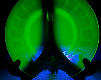 May include: A green glass plate with a scalloped edge, displayed on a black stand. The plate is translucent and has a slight green tint.