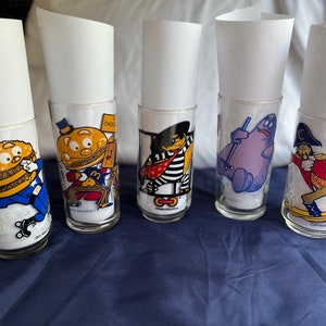 May include: Five vintage McDonald's drinking glasses featuring cartoon characters. Each clear glass has a different character design in vibrant colors, including a burger with a hat, a chef, and a purple character. White paper is inside each glass.