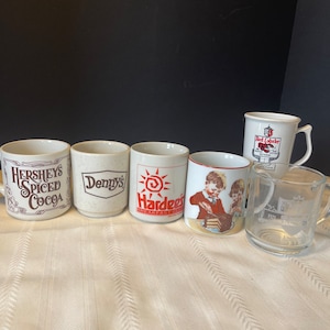 May include: Six ceramic mugs with various logos. The mugs are white with brown, red, and blue accents. The logos include Hershey's Spiced Cocoa, Denny's, Hardee's, Red Lobster, and a 1921 logo.