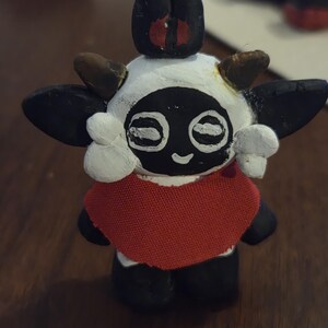 May include: A handcrafted figurine of a lamb-like character with a white head, black face, and large black ears. The figurine has a red cape and small brown horns. The character has a smiling expression and is standing upright.