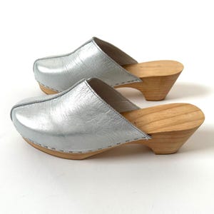 May include: A pair of silver leather clogs with wooden soles. The clogs have a rounded toe and a low, angled heel. The silver leather has a textured appearance. The wooden soles are light brown.