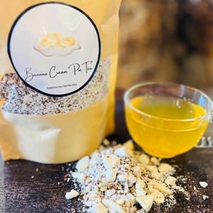 Könnte beinhalten: Ein brauner Papierbeutel mit einem weißen Etikett, auf dem "Banana Cream Pie Tea" steht und eine Zeichnung eines Bananencremekuchens abgebildet ist. Der Beutel ist mit losen Blättern gefüllt. Im Hintergrund steht eine Glas Tasse mit gelbem Tee.