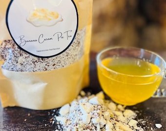 May include: A brown paper bag with a white label that says "Banana Cream Pie Tea" and a drawing of a banana cream pie. The bag is filled with loose leaf tea. A glass cup of yellow tea is in the background.