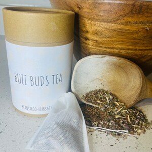 May include: A brown cardboard tube with a white label that says "Buzz Buds Tea" and "Beansxroo-Herbs&TF". A white tea bag is next to the tube, and a wooden spoon filled with loose leaf tea is in the background.