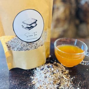 May include: A brown paper bag of S'mores Tea with a round label featuring a s'more illustration. A small pile of tea and a clear glass teacup filled with orange tea are next to the bag. The text on the label reads "S'mores Tea" and "beannaroo-herbsten".