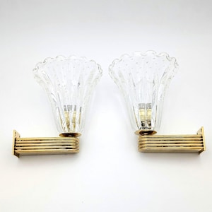 May include: A pair of wall sconces featuring clear, fluted glass shades with a textured, dotted pattern. Each shade is supported by a horizontal, multi-layered gold-toned metal arm with a rectangular mounting plate at the base for wall attachment.