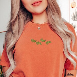 May include: A rust-colored t-shirt with a green embroidered design of four lizards. The shirt is loose-fitting and has a crew neck.