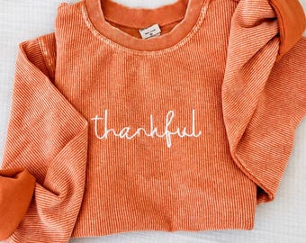 Op de afbeelding: Een sweatshirt met lange mouwen en ronde hals in een gebrande oranje kleur. Het kledingstuk is voorzien van het woord "thankful" geborduurd in wit cursief schrift in het midden van de borst. De stof heeft een verticaal geribbeld patroon.