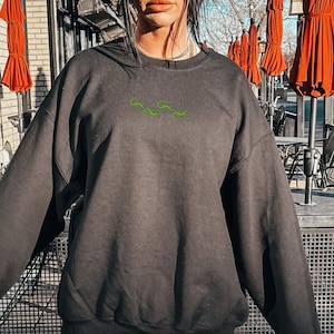 May include: A black sweatshirt with a green embroidered design. The sweatshirt is worn with black leggings and white sneakers.