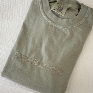Puede incluir: Camiseta verde salvia doblada con la palabra "bookworm" bordada en una fuente minimalista. La camiseta tiene una etiqueta Comfort Colors. Tambi&eacute;n es visible el texto "Pictured with Sandstone Shirt, Minimal Font, and Latte Thread".