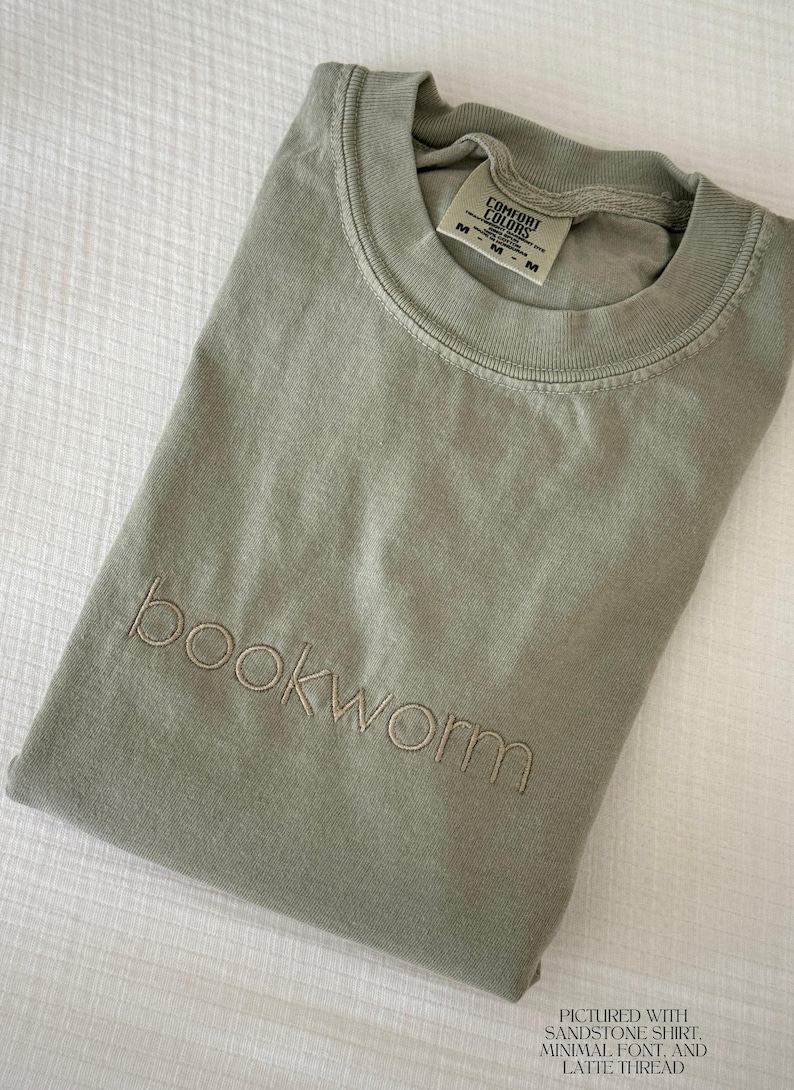 Puede incluir: Camiseta verde salvia doblada con la palabra "bookworm" bordada en una fuente minimalista. La camiseta tiene una etiqueta Comfort Colors. Tambi&eacute;n es visible el texto "Pictured with Sandstone Shirt, Minimal Font, and Latte Thread".