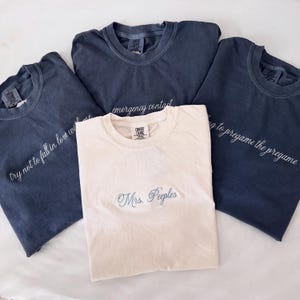 May include: Four t-shirts in shades of blue and cream. One cream-colored shirt has "Mrs. Peeples" embroidered in light blue. The blue shirts have white embroidered text: "try not to fall in love with me", "emergency contact", and "going to pregame the pregame".