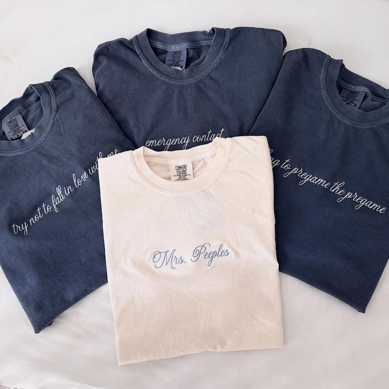 May include: Four t-shirts in shades of blue and cream. One cream-colored shirt has "Mrs. Peeples" embroidered in light blue. The blue shirts have white embroidered text: "try not to fall in love with me", "emergency contact", and "going to pregame the pregame".