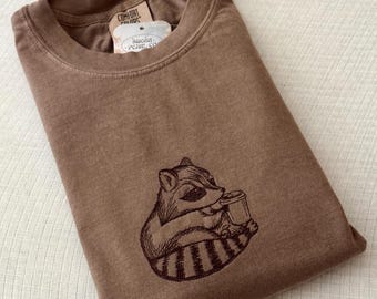 May include: Brown t-shirt with a raccoon graphic embroidered on the front. The raccoon is holding a cup. The shirt has a crew neck and a tag that says "Comfort Colors" and "Sugar Penny Co."