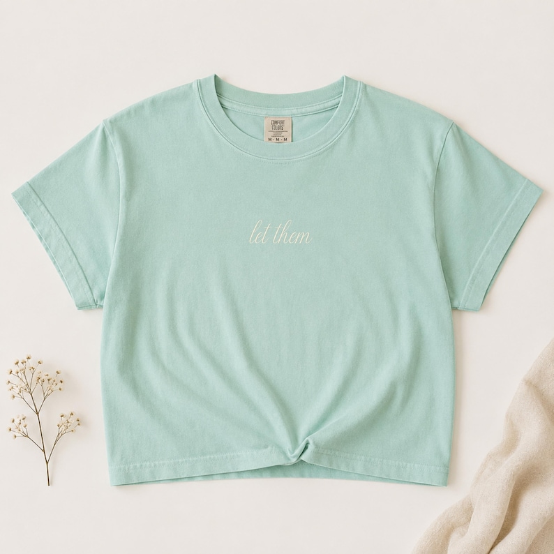 Puede incluir: Una camiseta de manga corta en color verde menta claro con cuello redondo. El centro del pecho presenta el texto "let them" en una fuente cursiva blanca. La etiqueta interior del cuello muestra la marca Comfort Colors y la talla M.