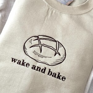 May include: A beige sweatshirt with a brown embroidered image of a loaf of bread and the text "wake and bake".