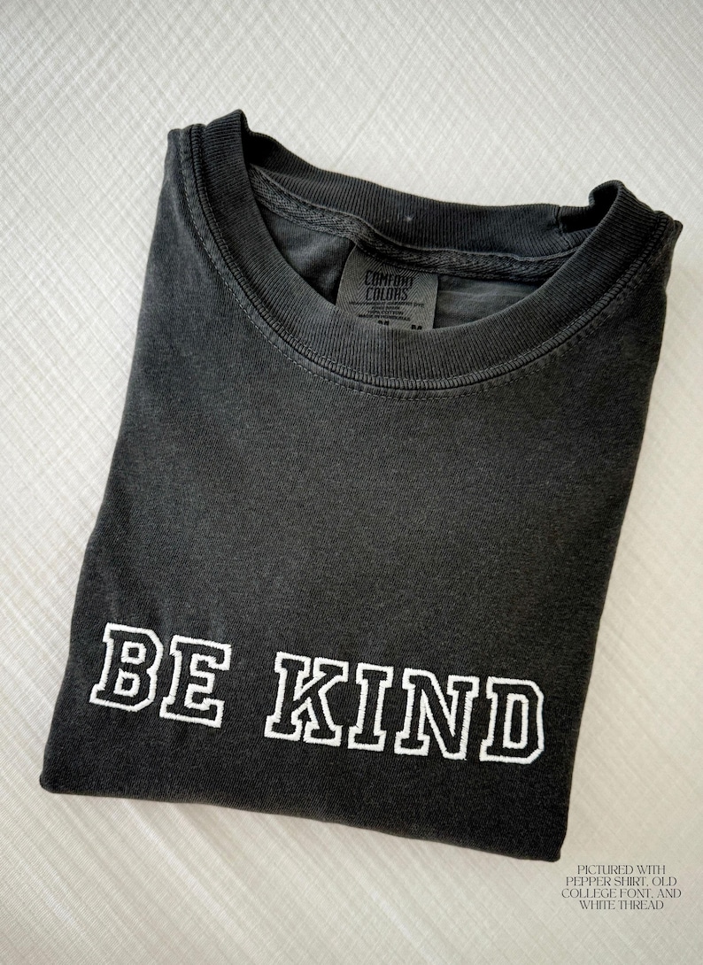 Puede incluir: Una camiseta gris oscuro doblada con las palabras "BE KIND" en blanco, con una fuente de estilo universitario. La camiseta tiene un aspecto vintage y una etiqueta Comfort Colors. El texto "PICTURED WITH PEPPER SHIRT, OLD COLLEGE FONT, AND WHITE THREAD" est&aacute; en la parte inferior.
