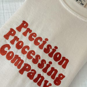 Puede incluir: Sudadera color crema con el texto bordado "Precision Processing Company" en una fuente naranja audaz. La sudadera tiene cuello redondo y una etiqueta que dice "Comfort Colors".
