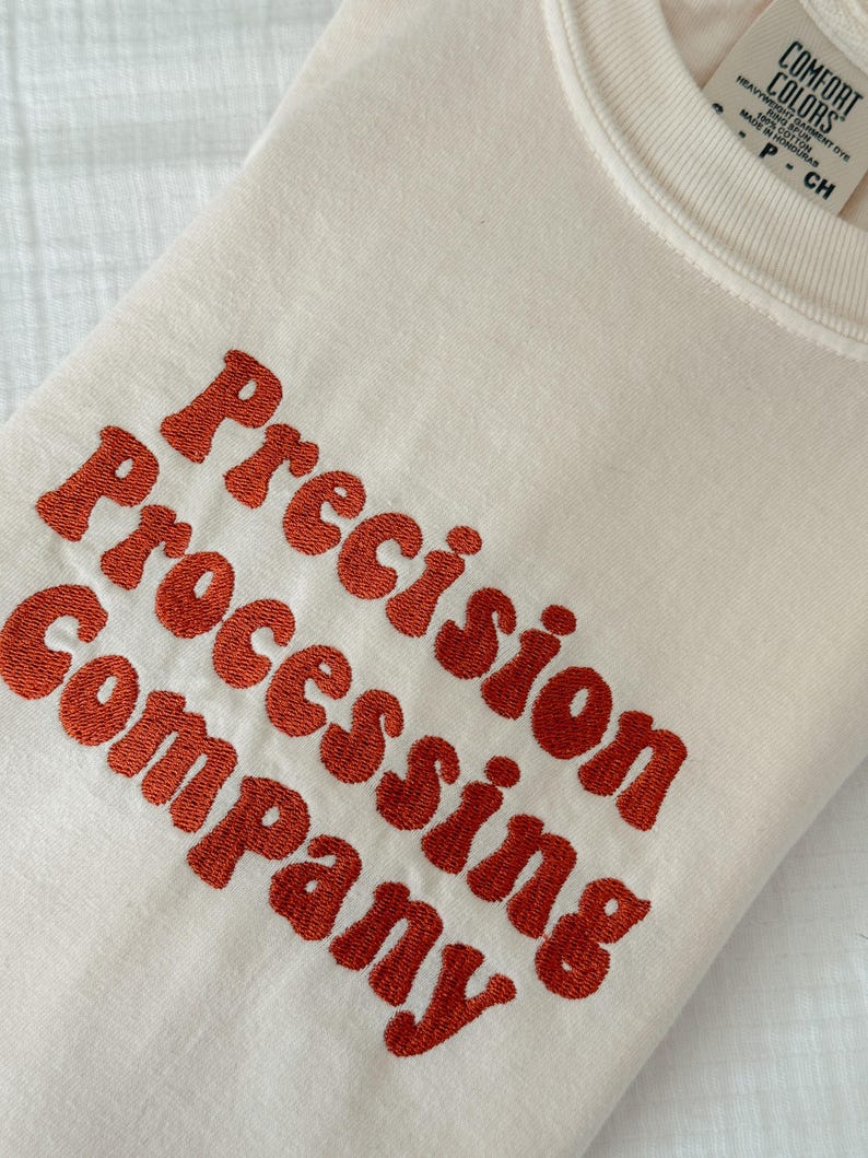Puede incluir: Sudadera color crema con el texto bordado "Precision Processing Company" en una fuente naranja audaz. La sudadera tiene cuello redondo y una etiqueta que dice "Comfort Colors".