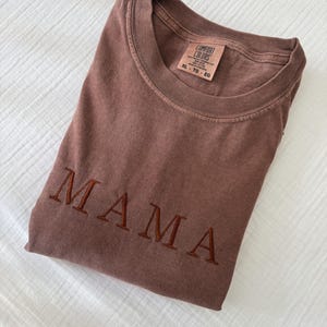 May include: A folded brown t-shirt with the embroidered word "MAMA" in a darker brown thread. The shirt has a crew neck and a tag that reads "COMFORT COLORS" and "XL - TG - EG". The text "PICTURED WITH CHOCOLATE THREAD" is at the bottom.