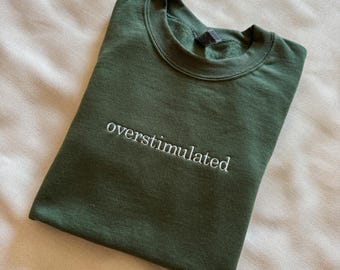 May include: A dark green sweatshirt with the word "overstimulated" embroidered in white on the front.